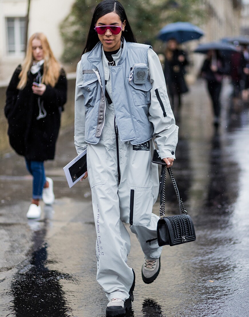 A street-styler wearing a grey jumpsuit, grey gilet, grey lace-up boots, a black Chanel bag and pink shades | ASOS Style Feed
