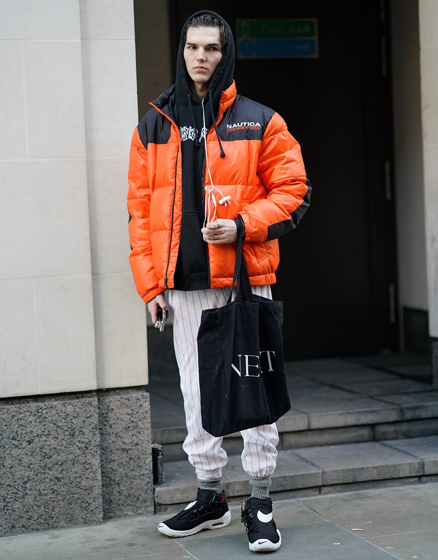 A street-styler wearing a black hoodie, striped trousers, orange puffer jacket and Nike trainers | ASOS Style Feed