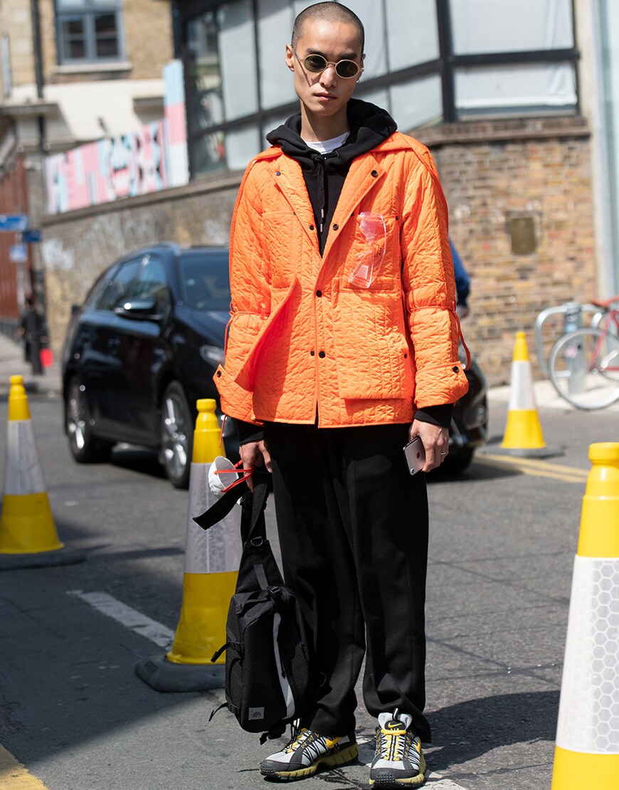 A street-styler wearing a black hoodie, shades, orange jacket, black trousers and trainers | ASOS Style Feed