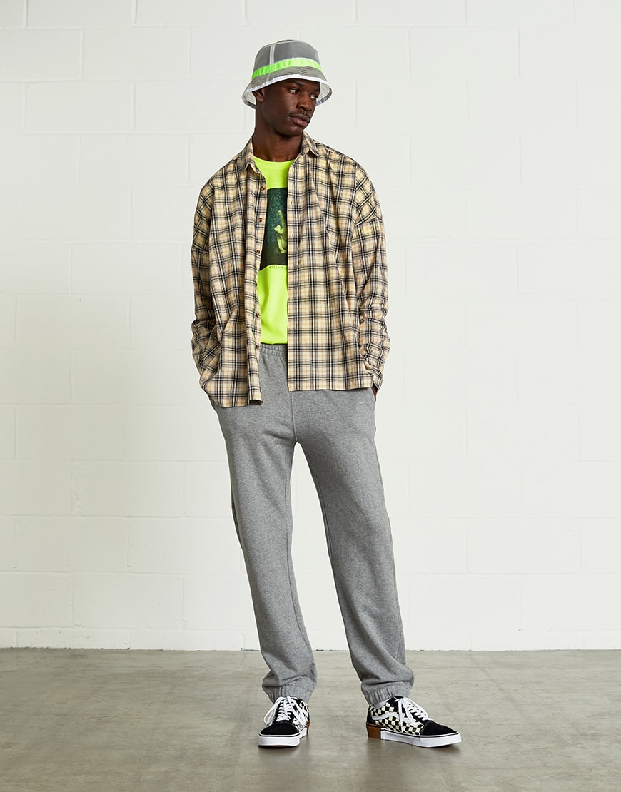 A picture of a man wearing a bucket hat, check shirt and a neon T-shirt with grey joggers.
