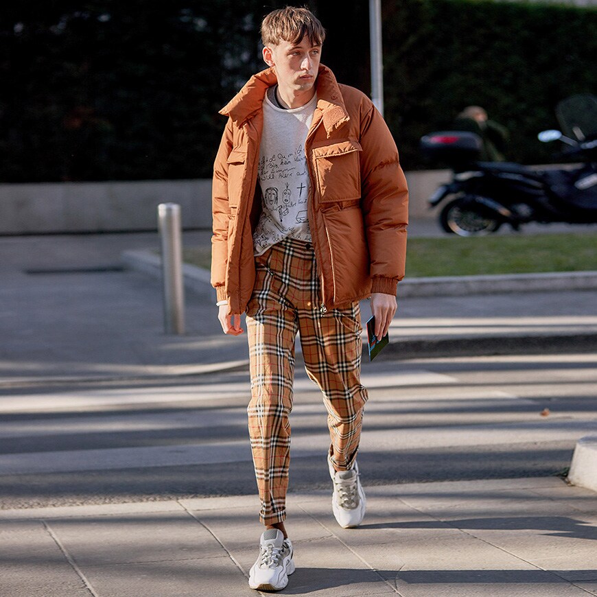 A picture of a man wearing a tan puffer jacket, white printed T-shirt, check trousers and white trainers.