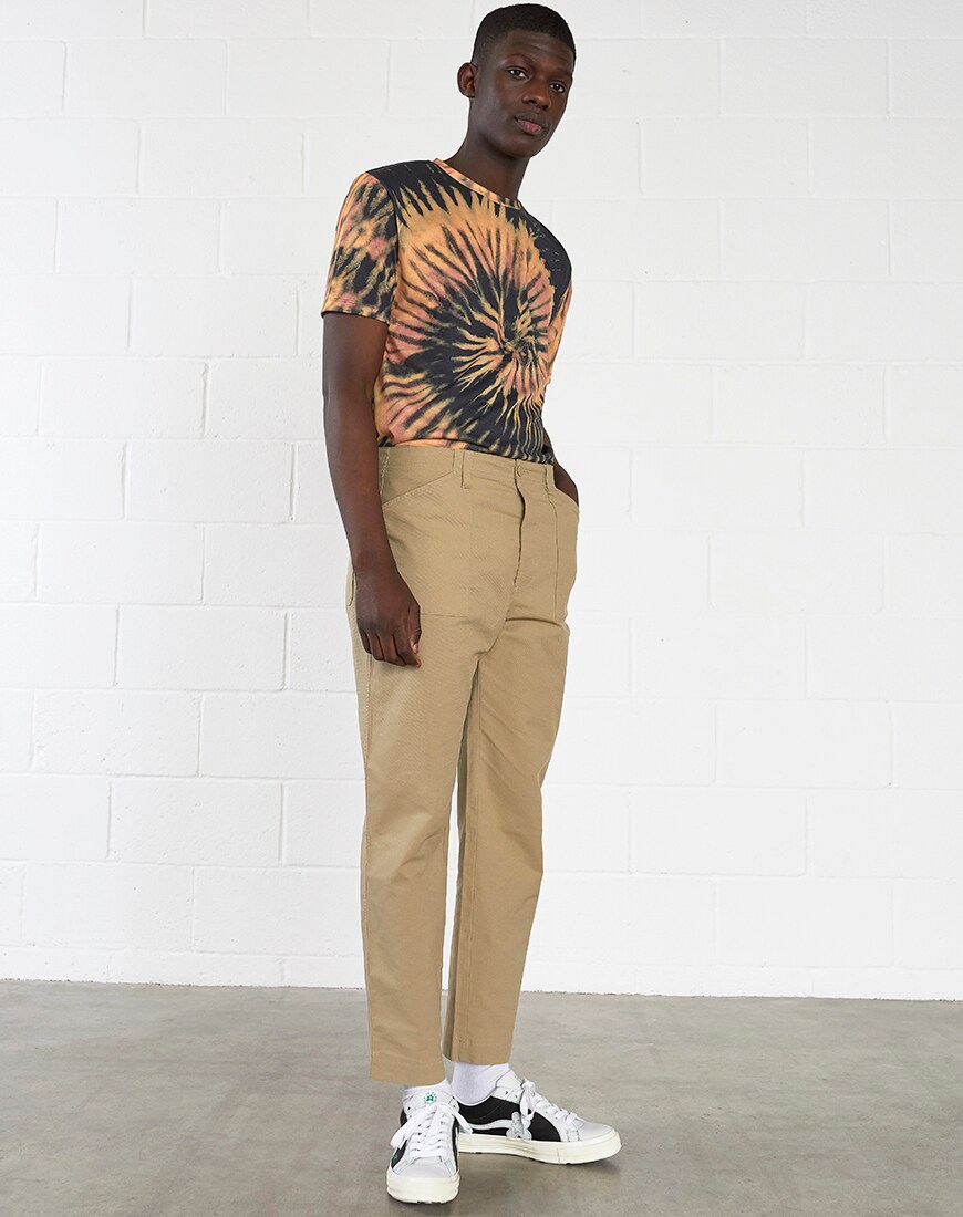 A picture f a model wearing a tie-dye T-shirt, camel-coloured trousers and Converse Golf Le fleur trainers. Available at ASOS.