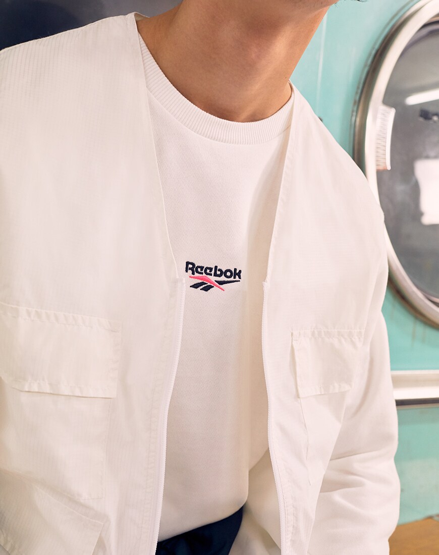A picture of a model wearing a Reebok T-shirt and a utility-style shirt. Available at ASOS.