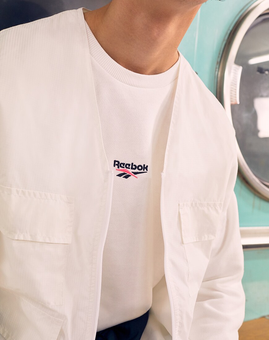 Une image d'un modèle portant un t-shirt Reebok et une chemise de style utilitaire. Disponible chez ASOS.