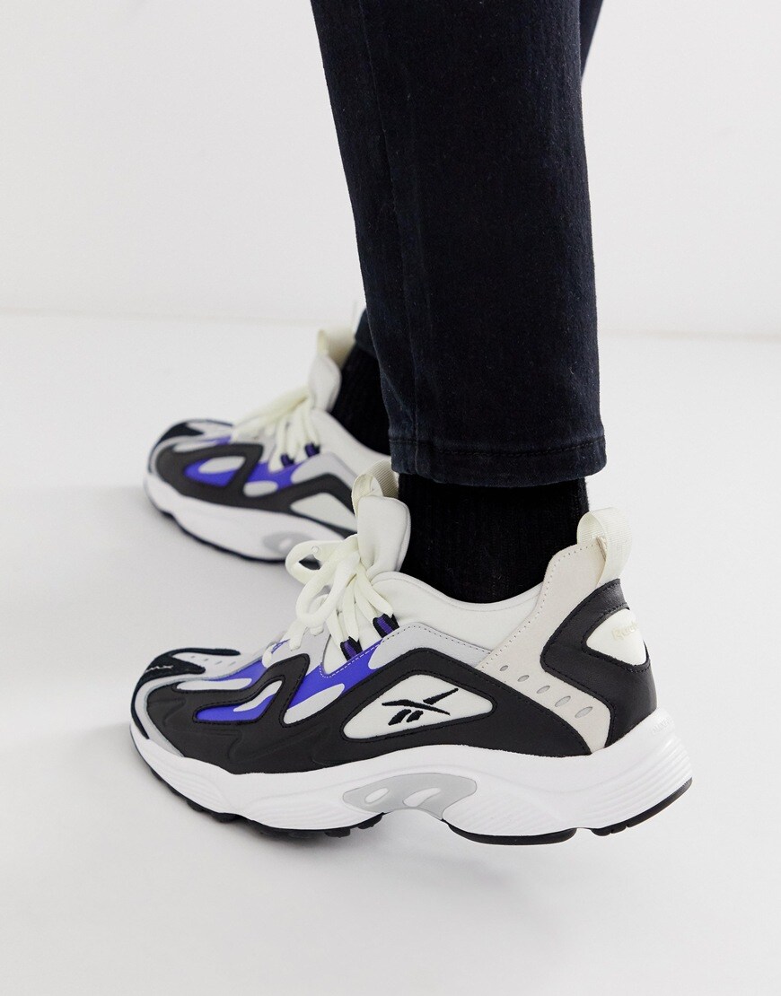 A picture of a model wearing a pair of Reebok DMX series 1200 trainers in a black, grey, blue and white colourway. Available at ASOS.