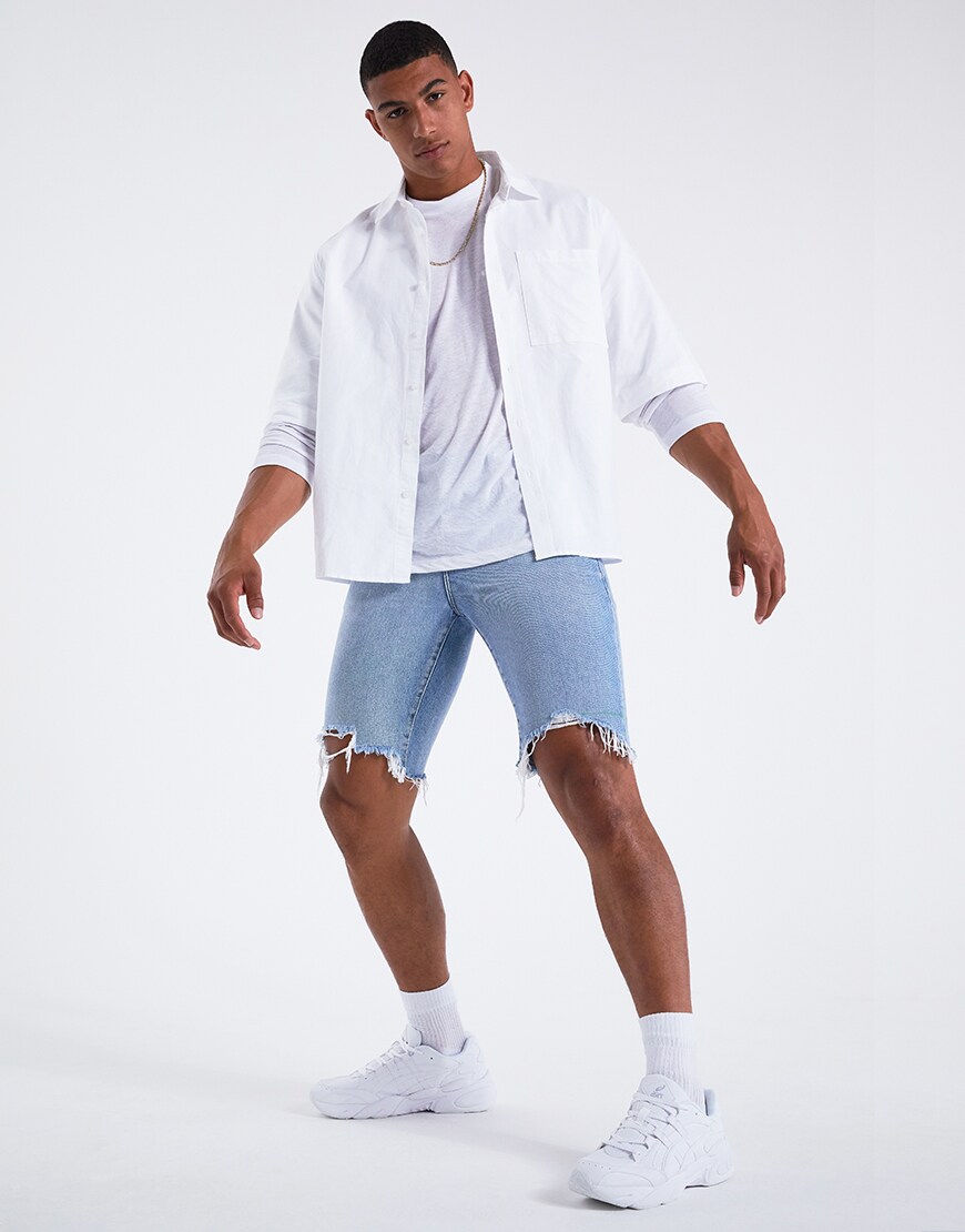 Image d'un modèle portant une chemise à manches courtes blanche sur un t-shirt à manches longues blanc, un short en jean déchiré et des baskets Asics. Disponible chez ASOS.