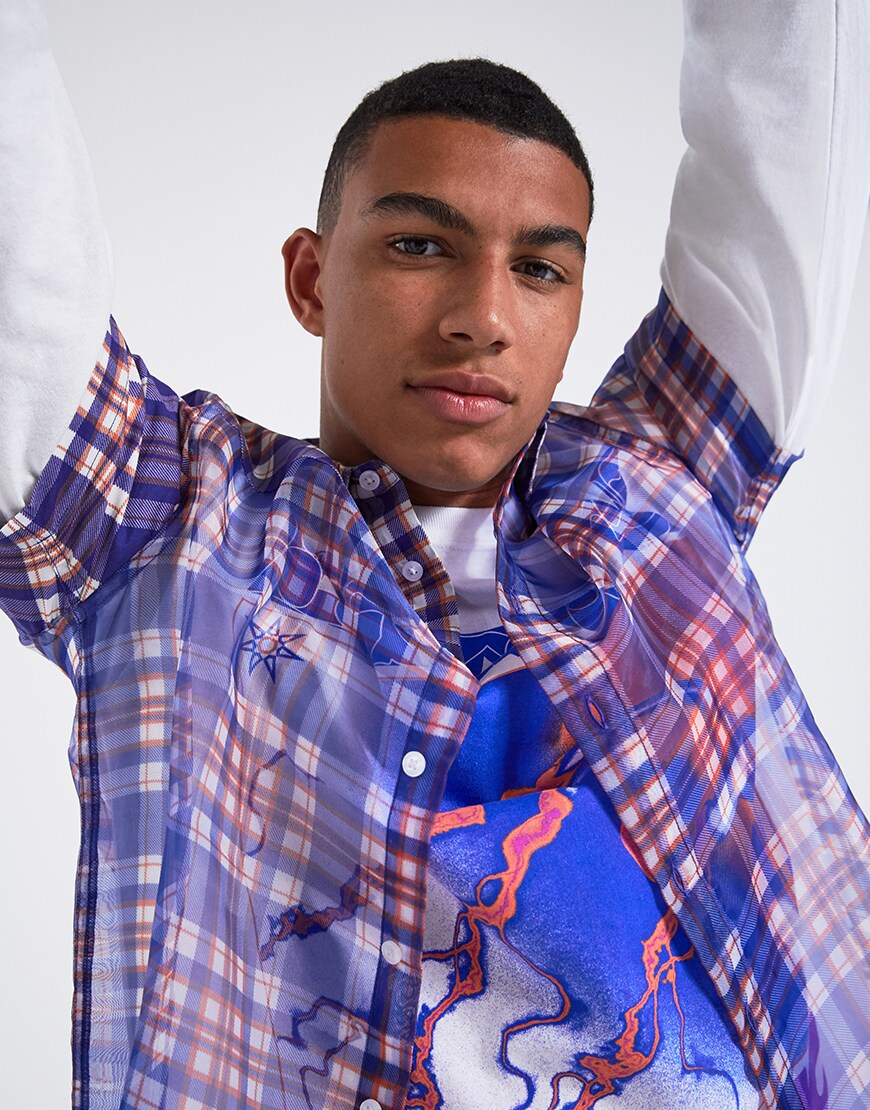 A picture of a model wearing a see-through check shirt over a long-sleeve printed T-shirt with ripped jeans and Nike trainers. Available at ASOS.