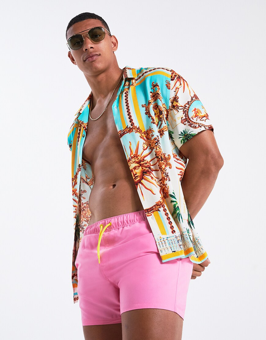 A picture of a model wearing a printed shirt, pink swim shorts and sliders. Available at ASOS