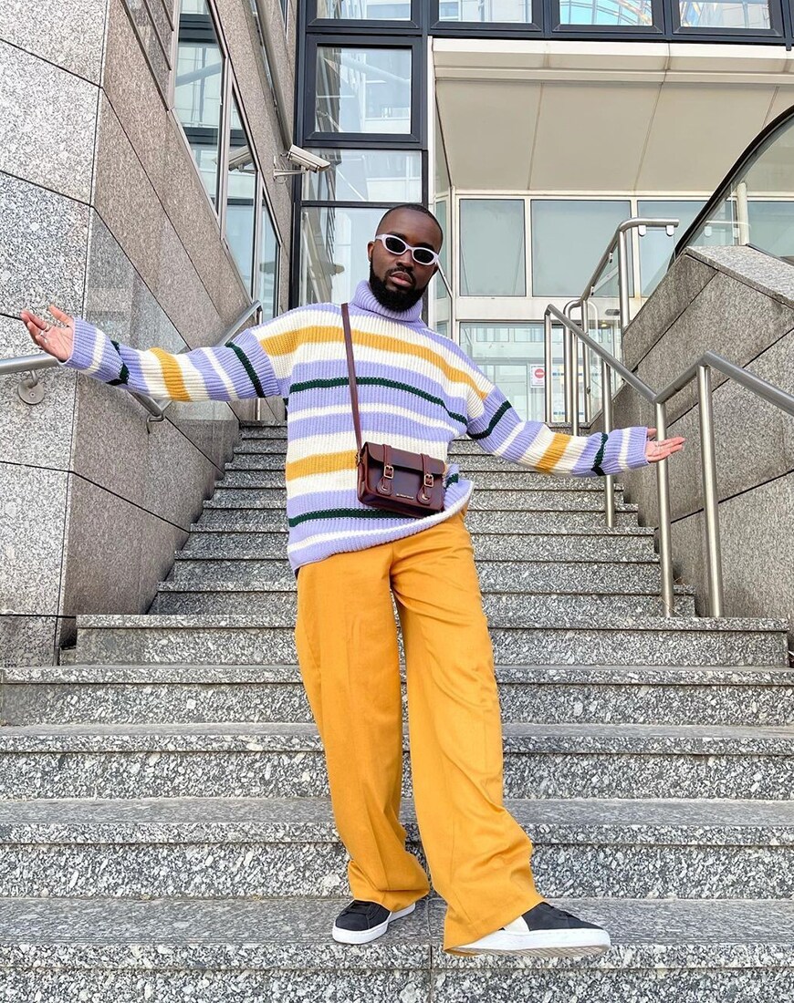 A man with his arms wide on concrete stairs while wearing a striped orange, purple and white jumper and black sunglasses | ASOS Style Feed 