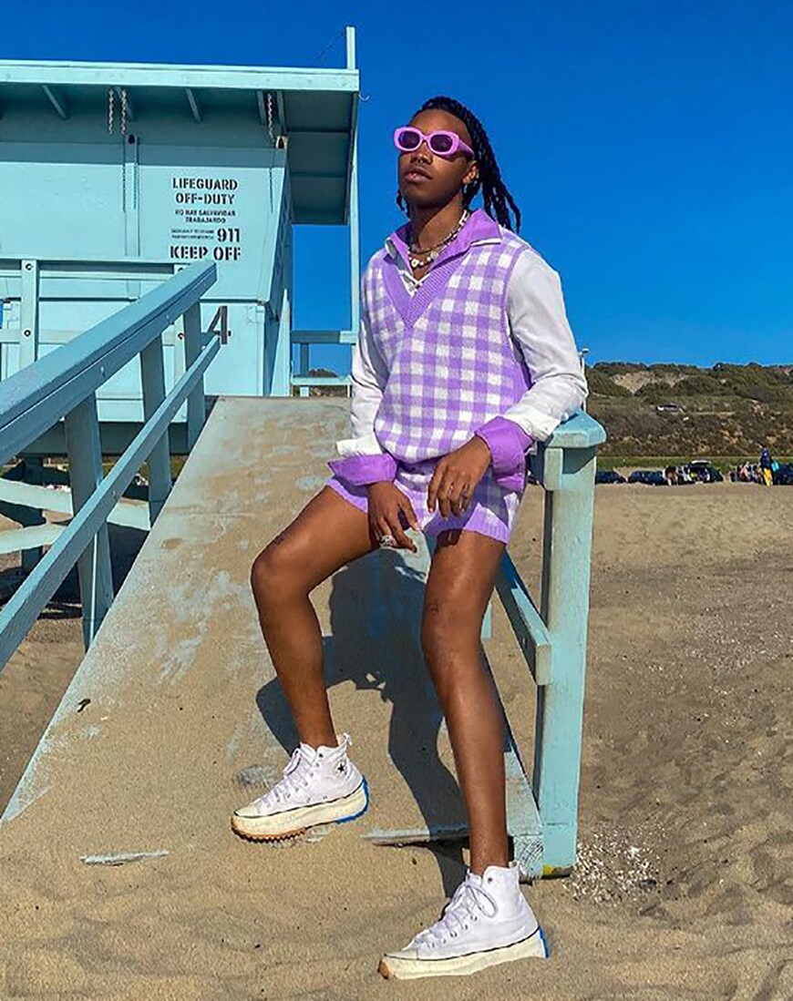 A man in a purple gingham sweater vest and matching shorts on a beach near a lifeguard station | ASOS Style Feed