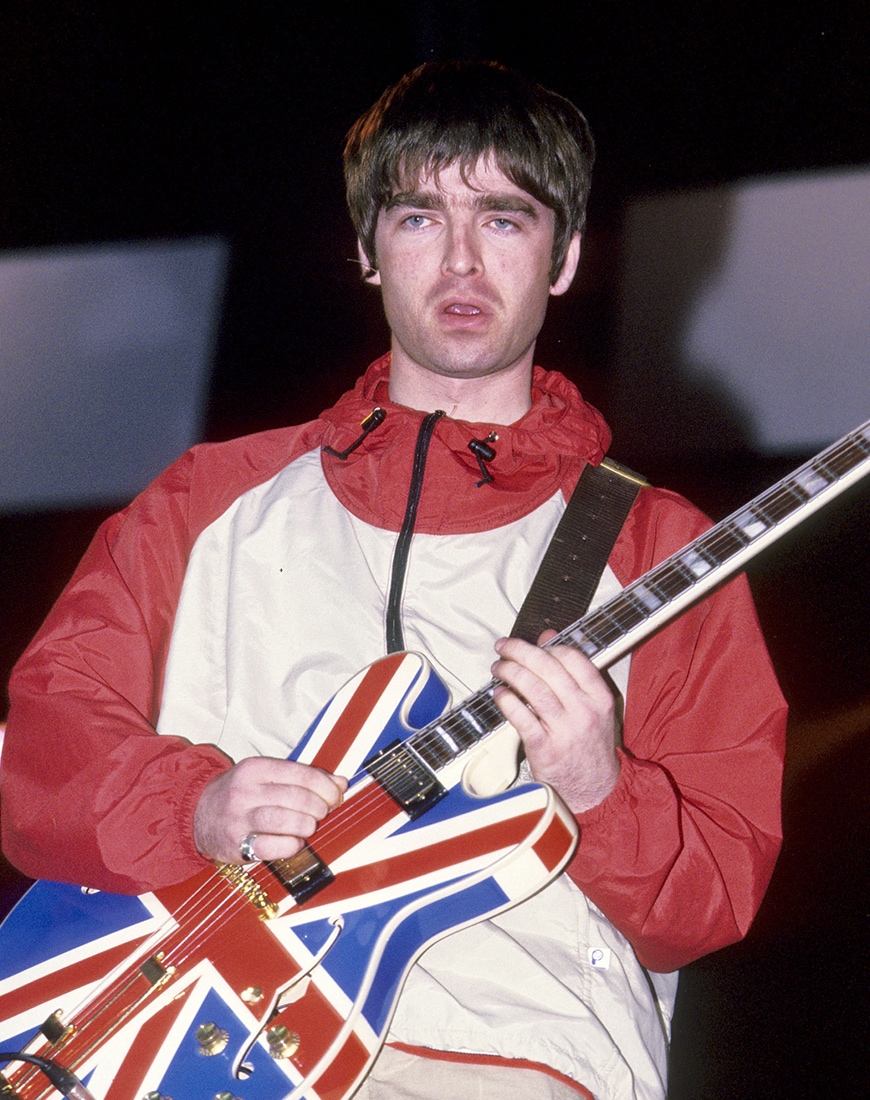 Noel Gallagher wearing red and white windbreaker jacket while holding Union Jack guitar