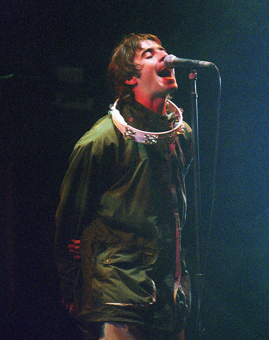 Liam Gallagher performing onstage while wearing a green parka jacket