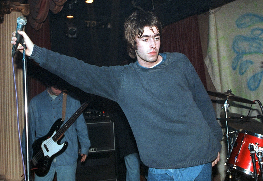 Liam Gallagher wearing a blue knitted jumper while holding a microphone