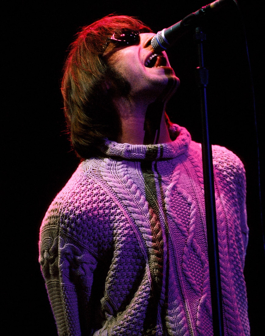 Liam Gallagher singing on stage wearing a cable knit jumper