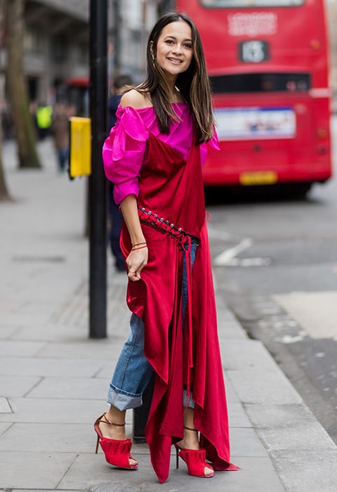 A fashion week attendee wearing a red silk slip dress with an off-the-shoulder pink shirt and cropped jeans.