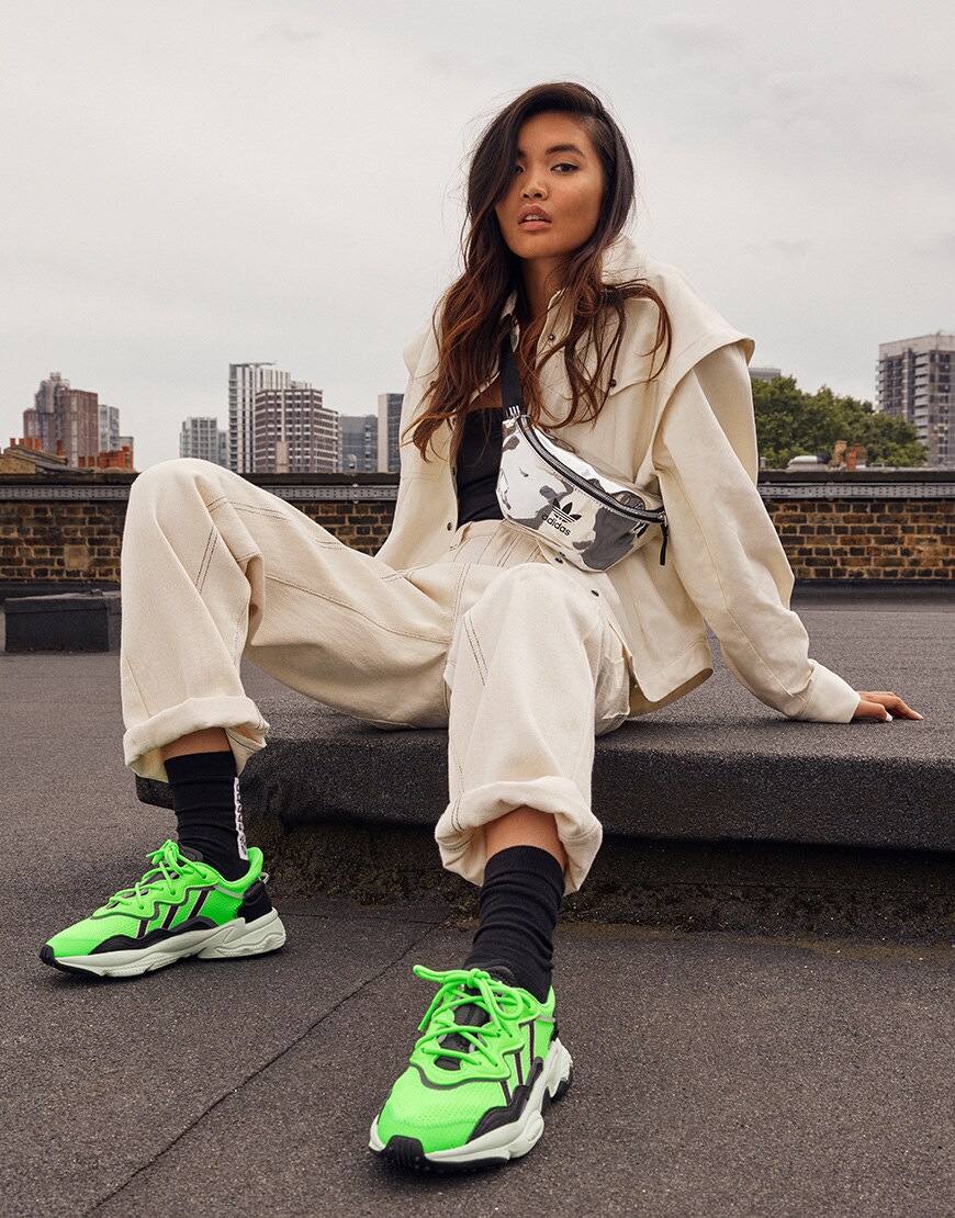 A picture of a model wearing a pair of neon-green adidas Ozweego trainers. Available at ASOS.