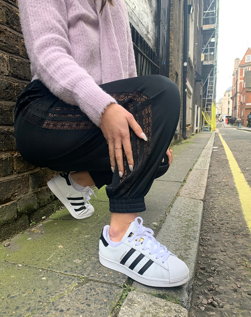 A picture of an ASOS Insider, ASOS Olive, wearing a pink fluffy cardigan with black adidas tracksuit bottoms and white adidas Superstar trainers.