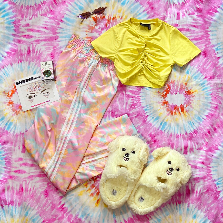A picture of a festival outfit against a pink tie-dye backgorund. The outfit consists of a yellow crop top, tie-dye adidas joggers, face and body gems and novelty puppy slippers. Available at ASOS.