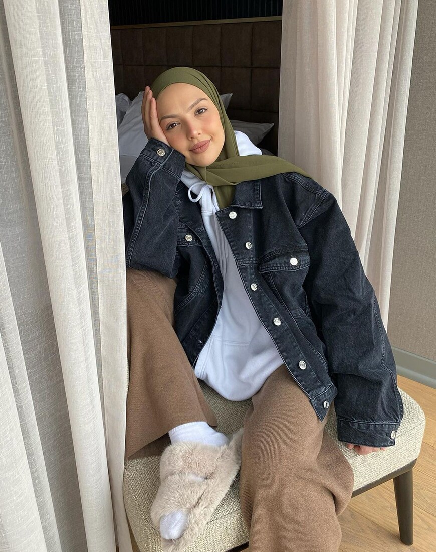 A woman wearing a headscarf, black denim jacket and taupe knit trousers while sitting near flowy curtains | ASOS Style Feed