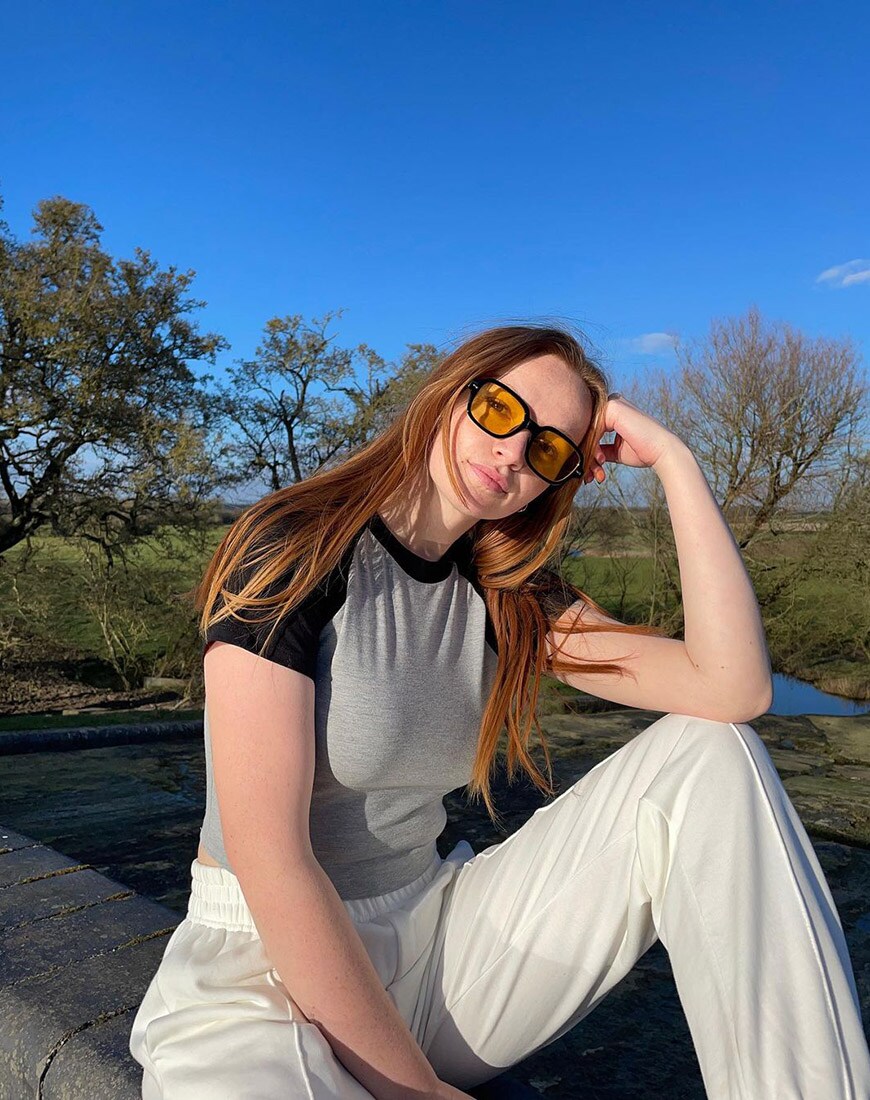 ASOS Scarlett sitting in a grey baseball tee and white joggers in front of trees and a blue sky | ASOS Style Feed
