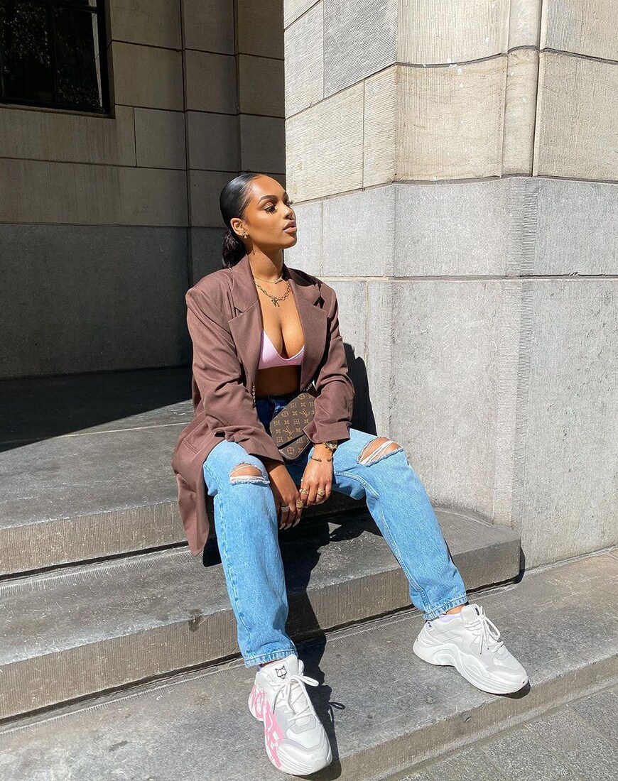 A woman sits on concrete steps wearing a brown dad blazer, pink bralette and blue ripped jeans | ASOS Style Feed