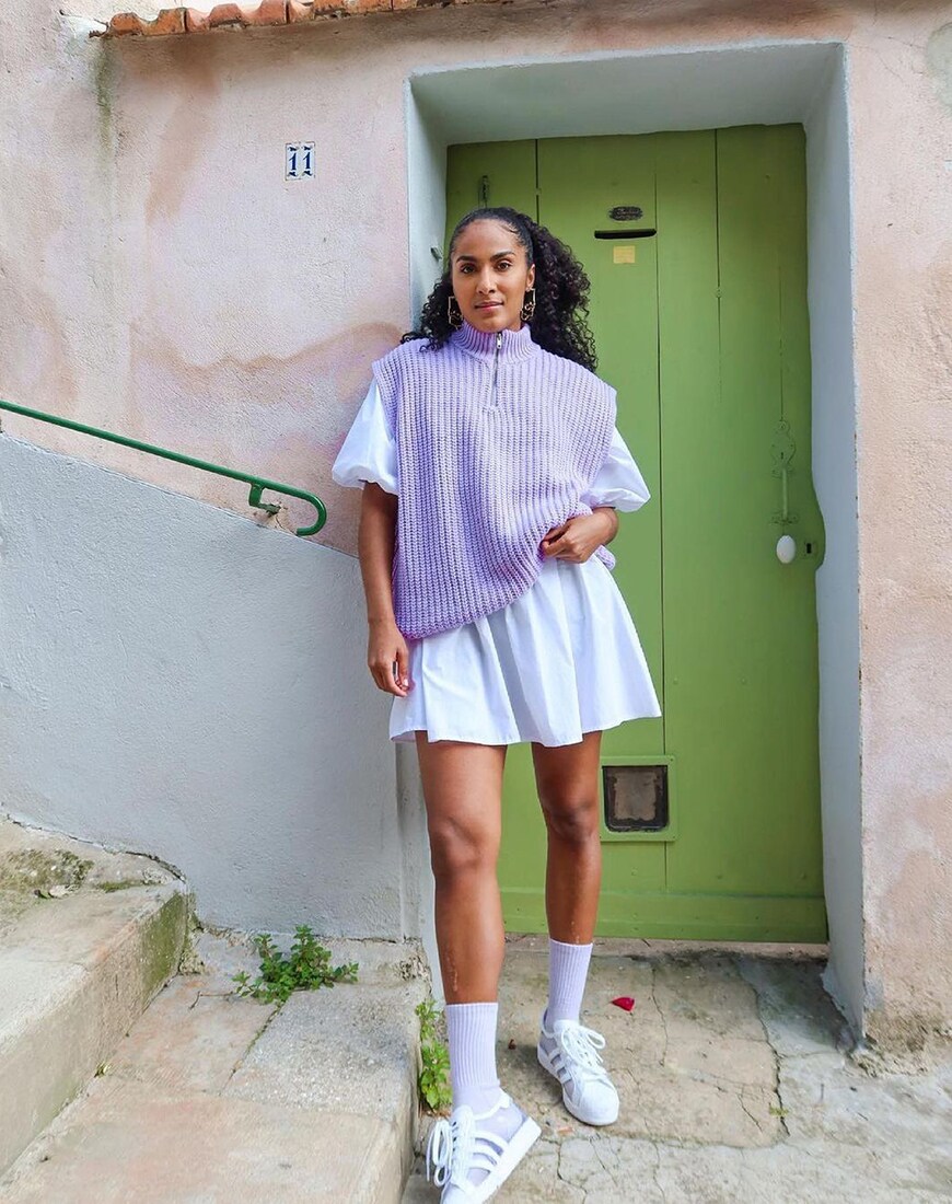 A woman with long curly hair standing in front of a green door with a purple knit vest over a white tee shirt and white tennis skirt | ASOS Style Feed