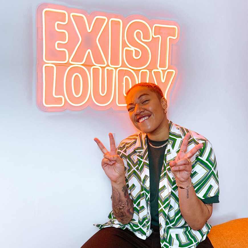 Tanya Compas standing in front of an 'Exist Loudly' neon sign