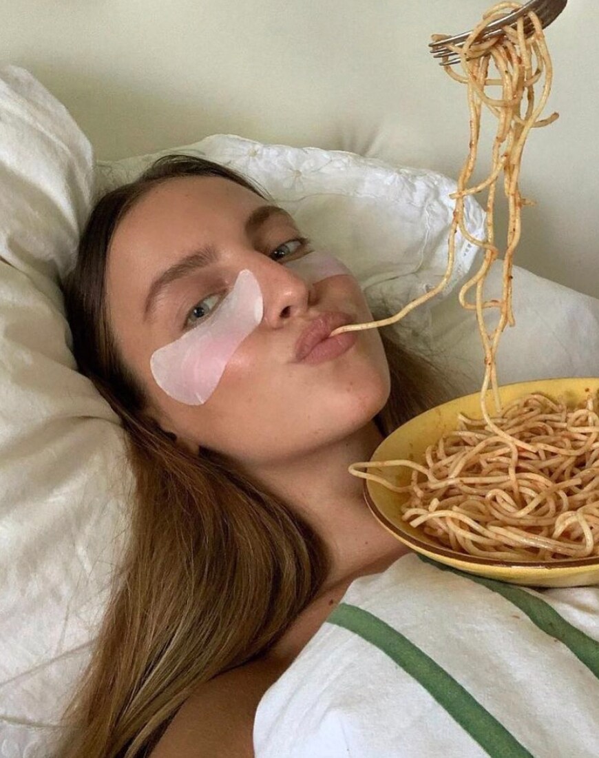 A woman eating spaghetti in bed with under-eye masks on