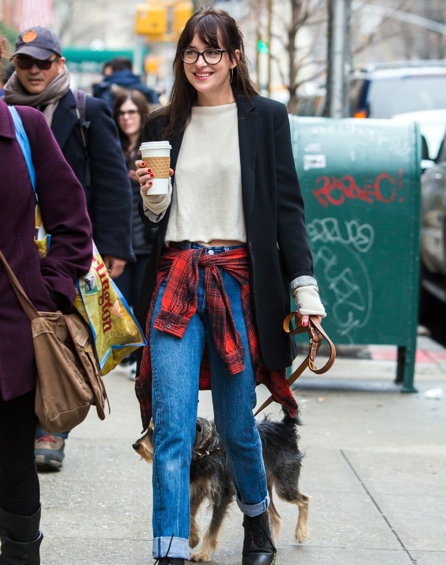 Dakota Johnson is on a dog walk, holding a coffee and wearing blue jeans, DMs, a white jumper and a black blazer. She has a red plaid shirt tied around her waist.| ASOS Style Feed