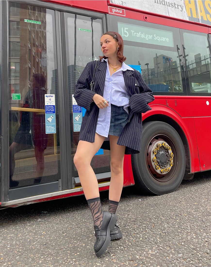 A woman stands in front of a bus wearing a navy blue pinstripe blazer, blue denim skirt, oversized shirt and loafers | ASOS Style Feed