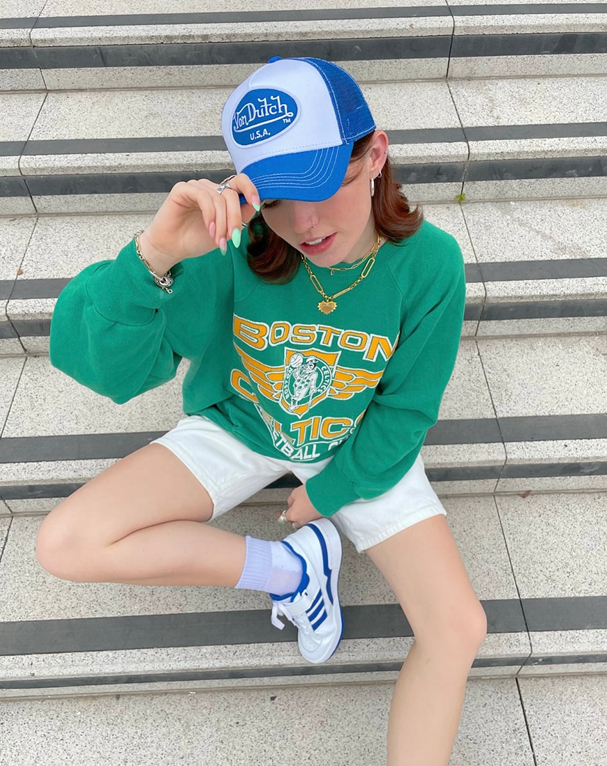 A woman sits on concrete steps wearing a blue and white trucker hat, oversized green sweatshirt and white shorts | ASOS Style Feed