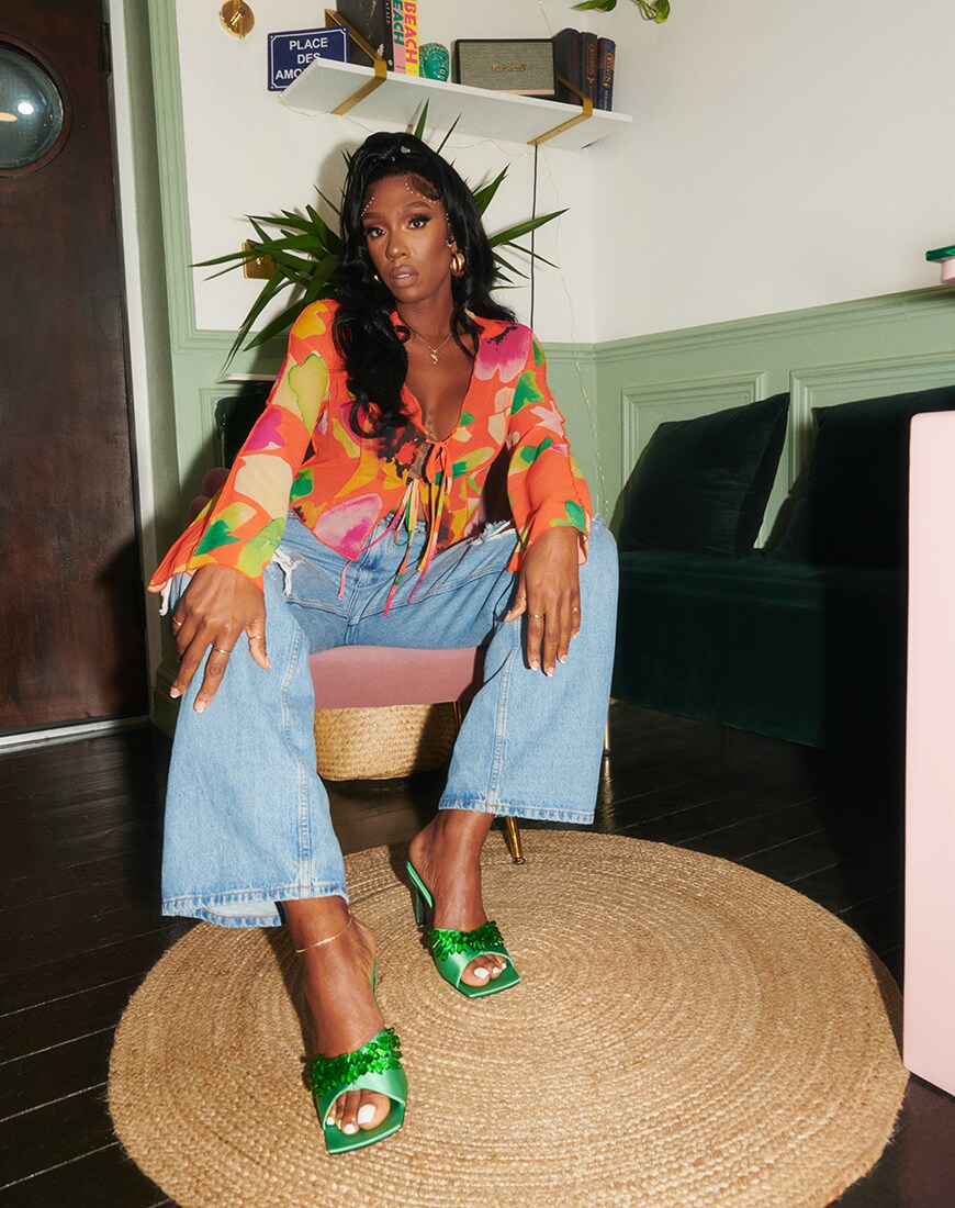 Irene Agbontaen sitting on a chair facing the camera, wearing blue flared jeans, green TTYA mules and a red and orange floral top | ASOS Style Feed
