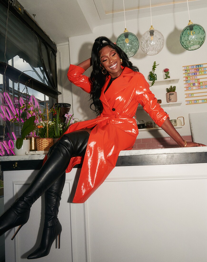Irene sits on a counter top with black TTYA thigh-high boots and an orange PU trench coat | ASOS Style Feed