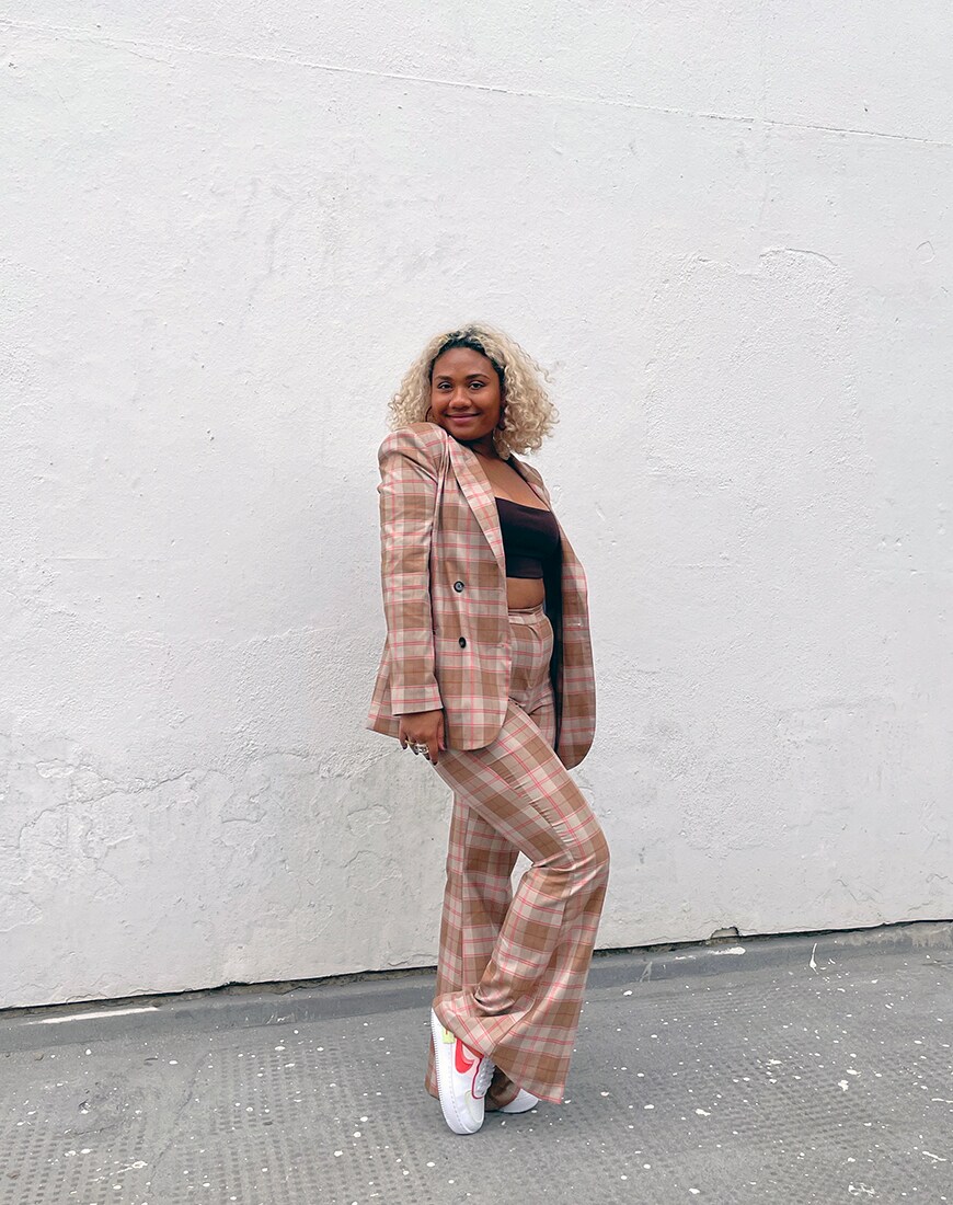 Ebony stands against a white wall in a pink and brown plaid suit, with a brown crop top and white and pink Nike trainers | ASOS Style Feed