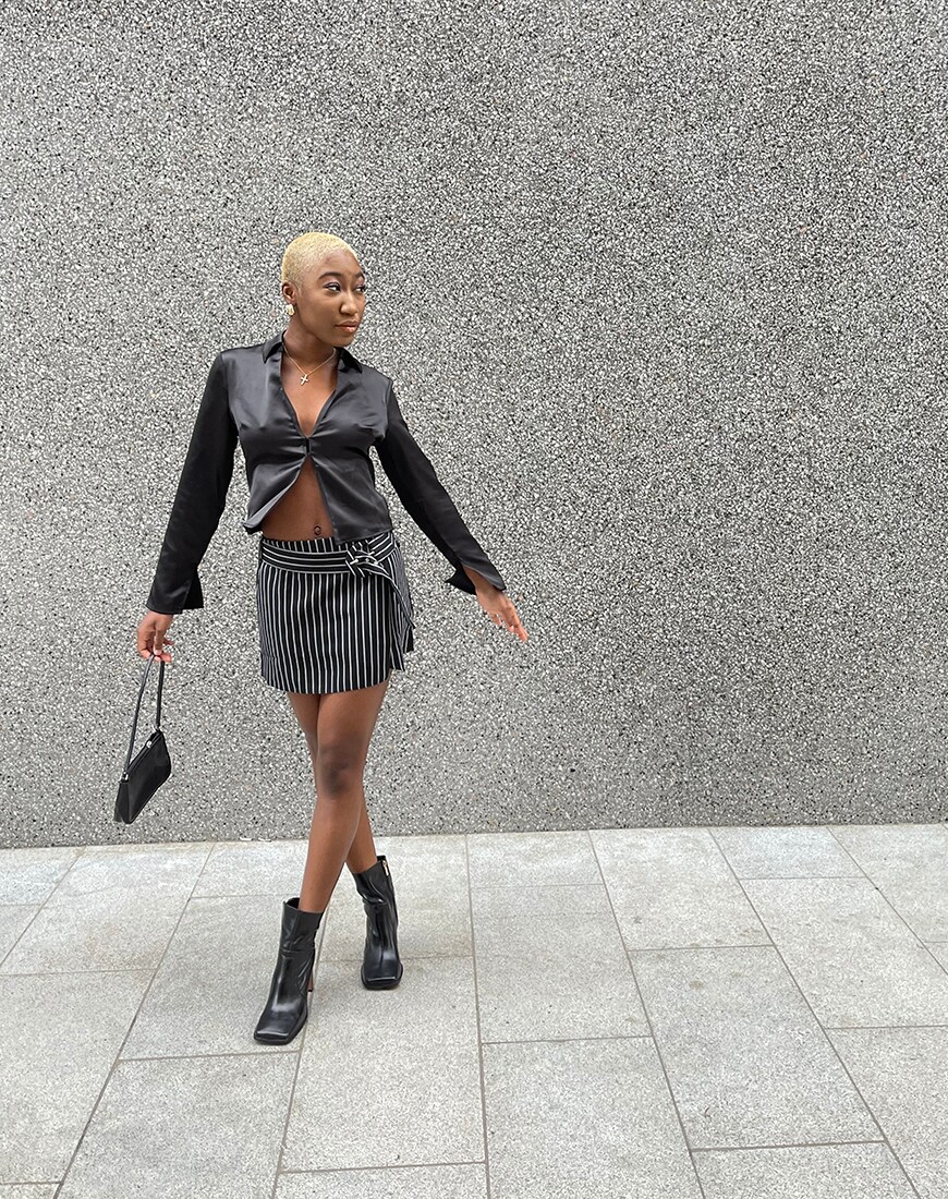Kamaria stands against a grey wall in a black blouse with a single button, black mini skirt and black heeled boots | ASOS Style Feed