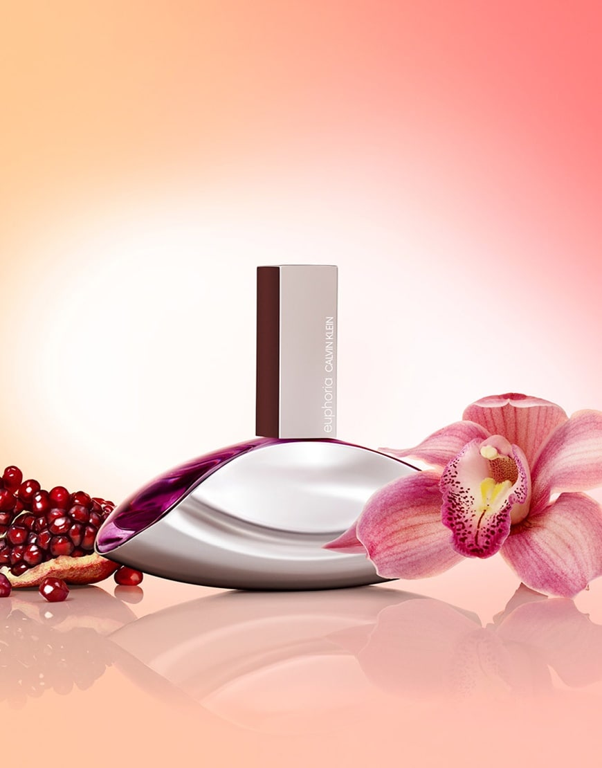 A picture of the Calvin Klein Euphoria perfume against a pink and orange background. Behind the perfume you can see a pomegranate and in the foreground there is an orchid. | ASOS Style Feed