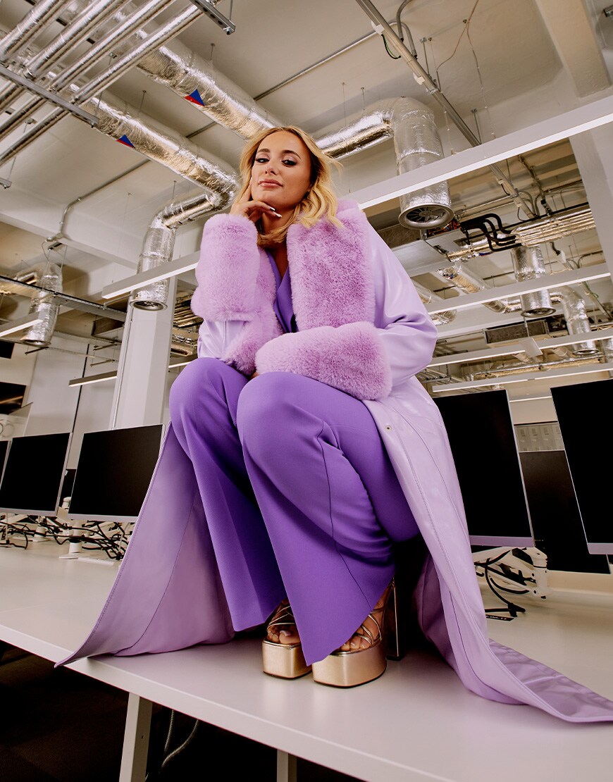 Millie crouches on top of a work desk in a head-to-toe lilac outfit – a long tench coat, wide leg trousers and a shirt | ASOS