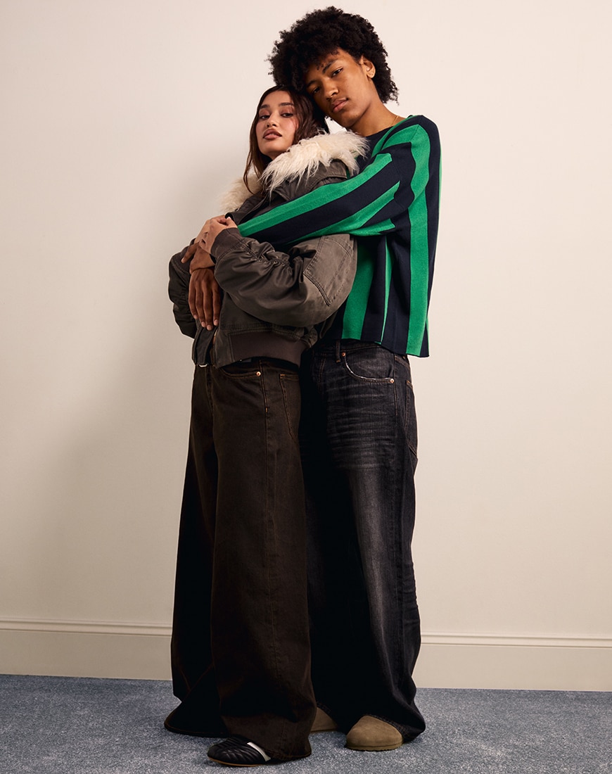 Woman wearing baggy jacket with grey jeans with man wearing green and black striped t shirt and black jeans