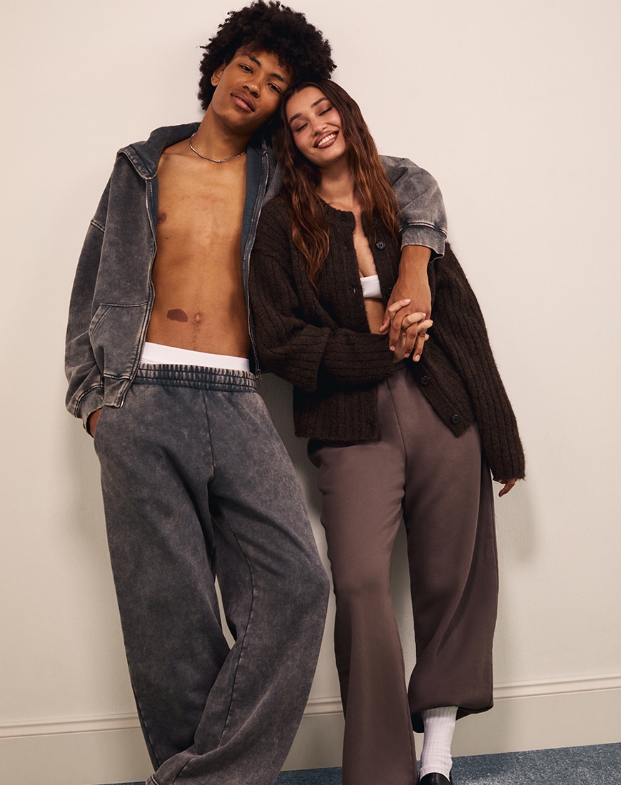 Man wearing acid wash grey tracksuit with his arm around woman wearing brown knit cardigan with brown joggers