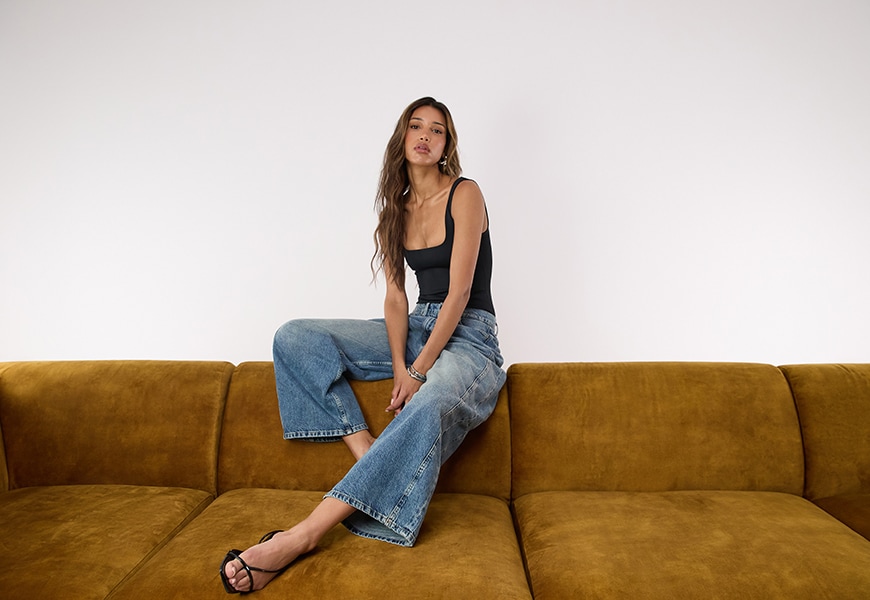 Model wearing a black top and jeans while sitting atop a sofa